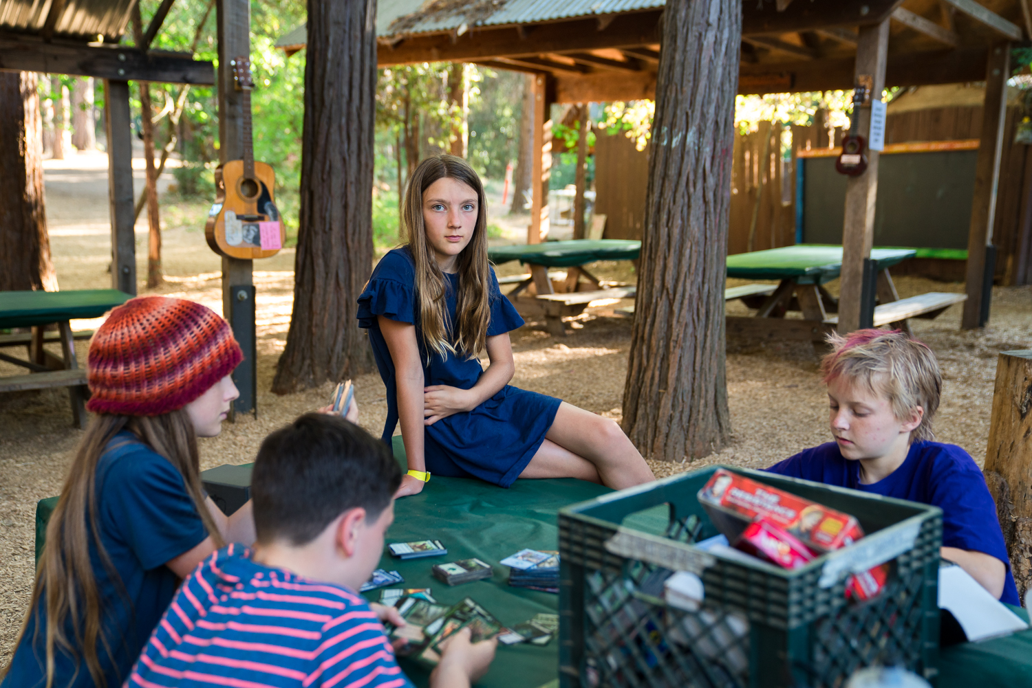 A girl sits on a picnic table where three boys are playing a card game.