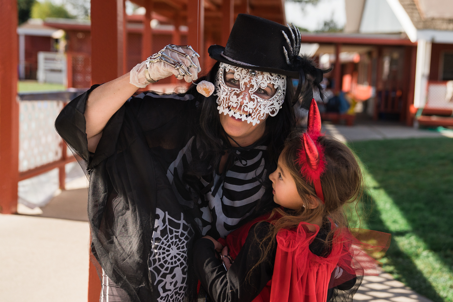 A woman in a lace skeleton mask and skeleton costume helps a young girl dressed like a devil to reach a donut hole hanging on a string.