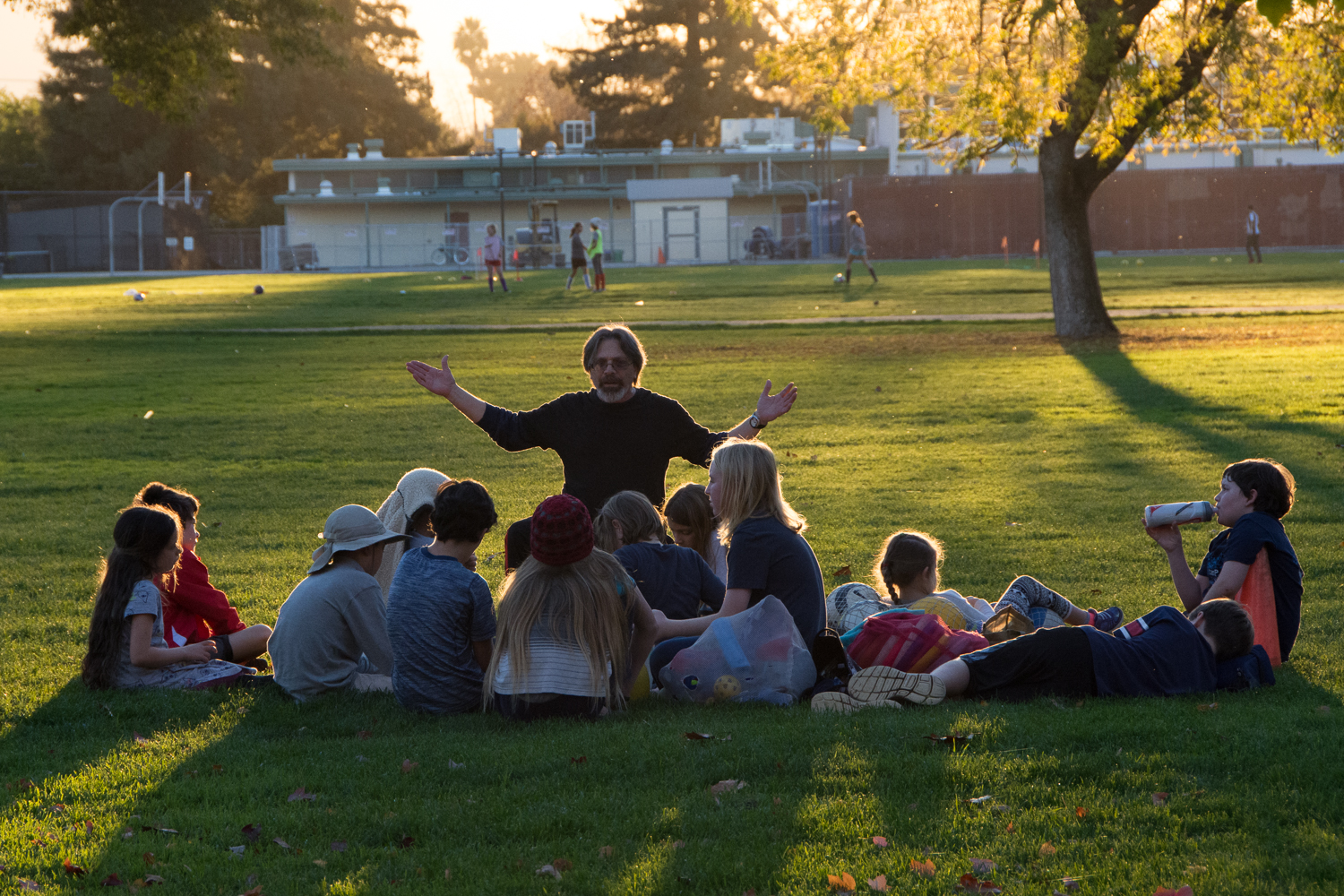 Man with arms raised speaks to a group of children sitting on the grass at dusk.